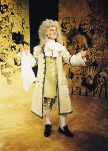The author playing a fop in a restoration comedy big wig, long cream jacket, black heels, long white hanky against a yellow background