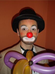 Clown. Red nose, white mouth, black hat, tan jacket, purple balloon flower