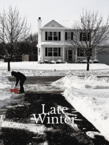 Snowy driveway. Man in the distance wearing winter clothes shoveling. Mostly black and white. Shovel is orange.